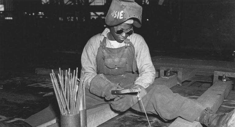 Black woman welder, glasses on, visor up, sits straddling her work, torch in hand