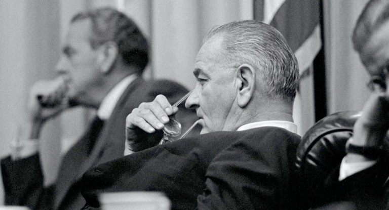 Profile of President Lyndon Johnson leaning back in a chair with one arm of the frame of his glasses in his mouth.