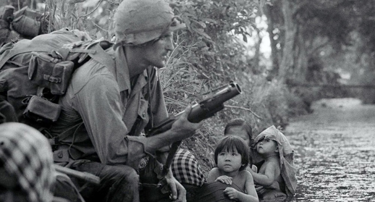 A soldier crouches on the banks of a stream alongside a Vietnamese family.