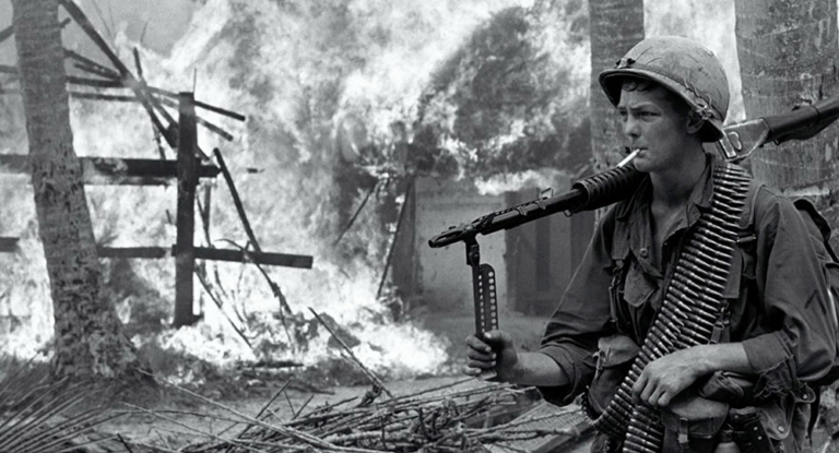 An American soldier stands beside a burning Vietnamese village.